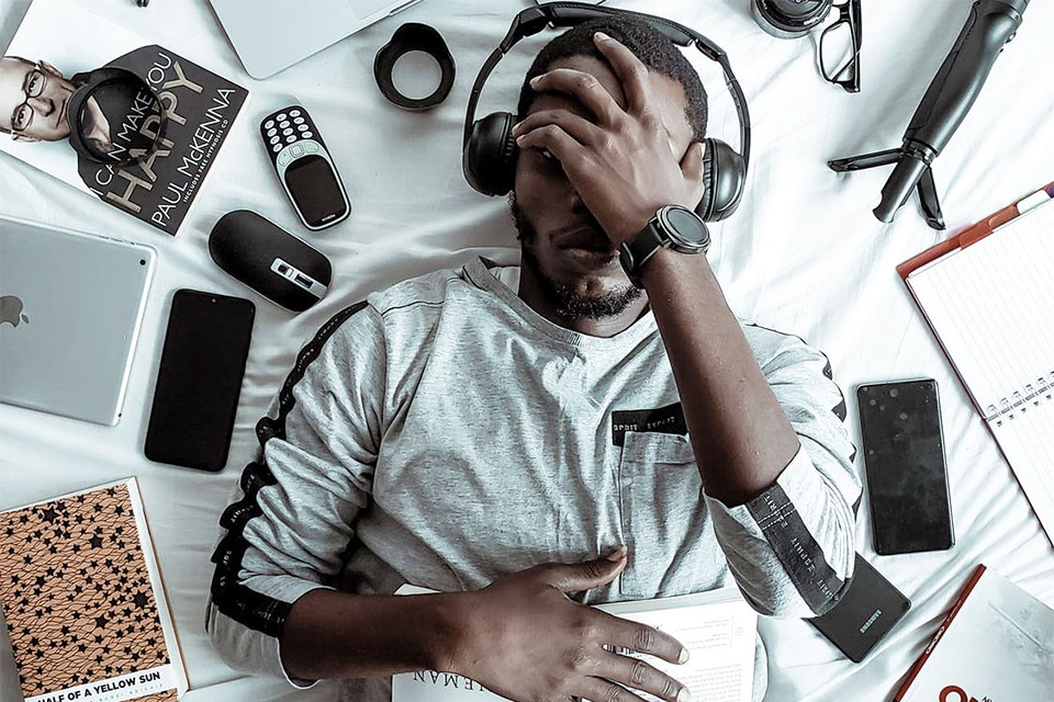 man lying in bed tired with laptop, cell phone and books