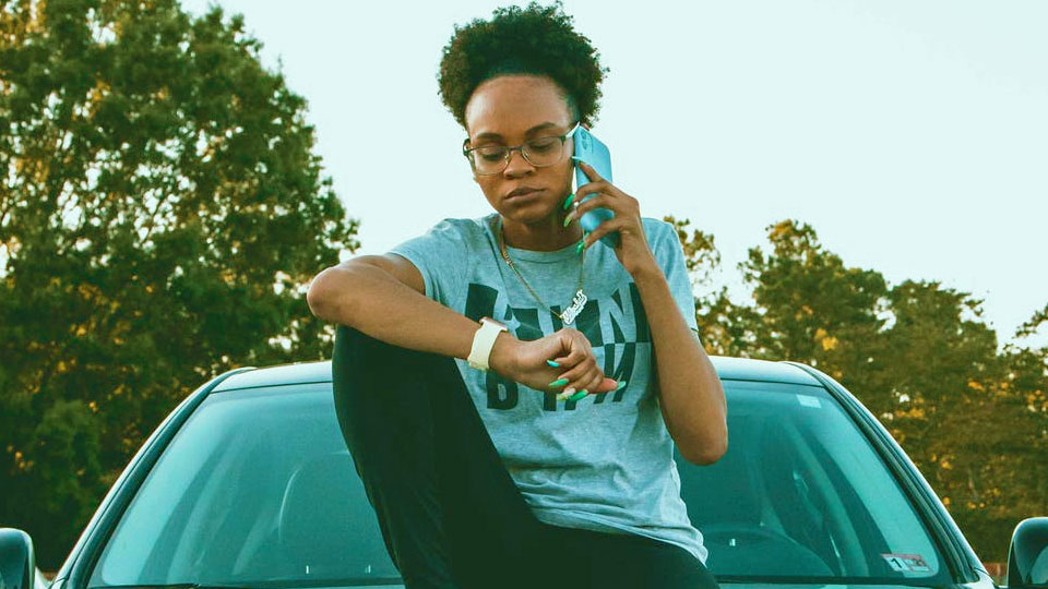woman sitting on the hood of a car with a cell phone and looking at her watch.