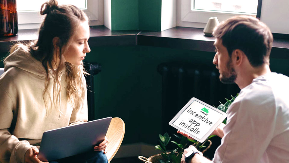 a woman with laptop on her lap looking at the android tablet that a man is holding