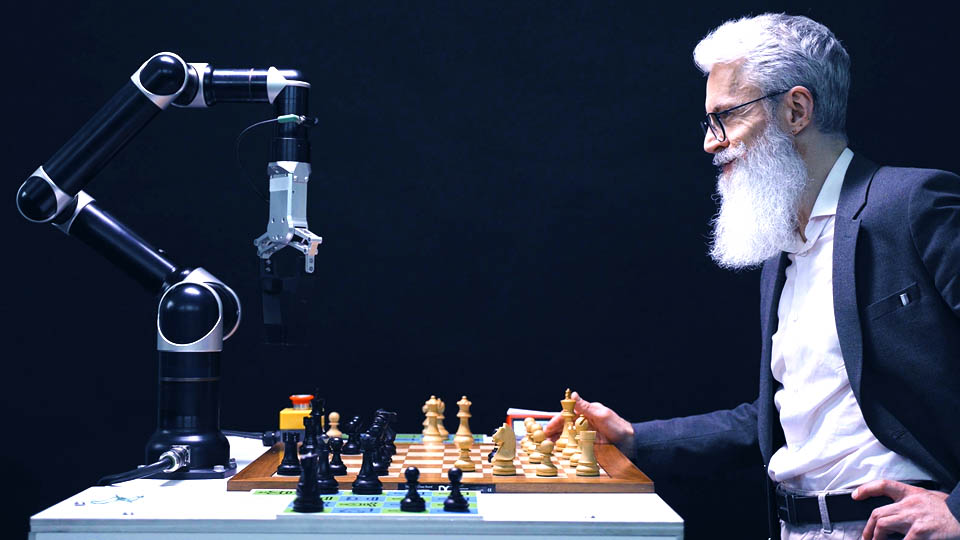 a bearded man playing chess with a robot arm