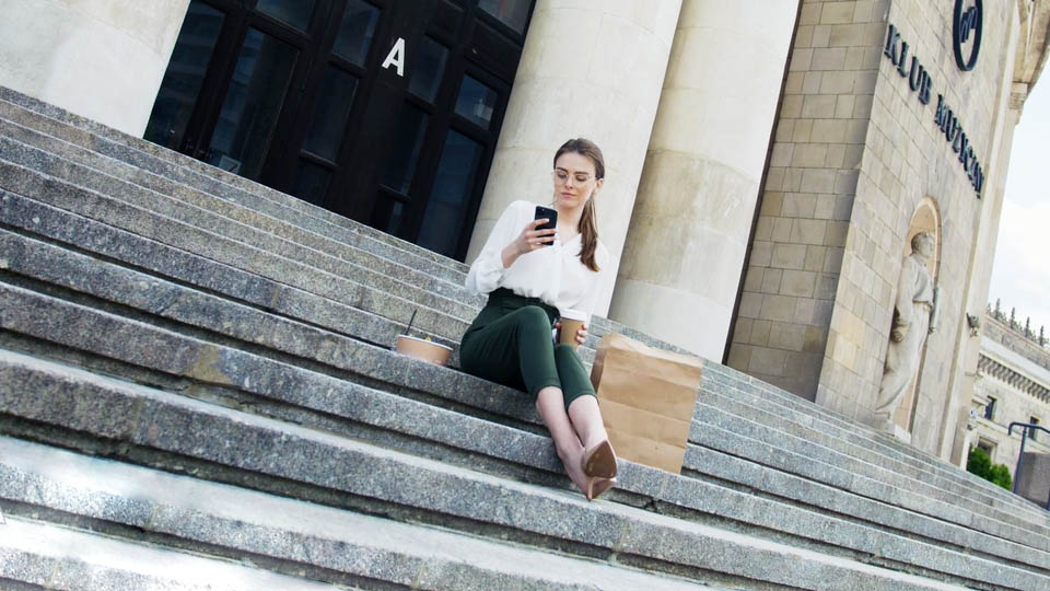 woman sitting holding a smartphone and a coffee in front of the congress hall in warsaw