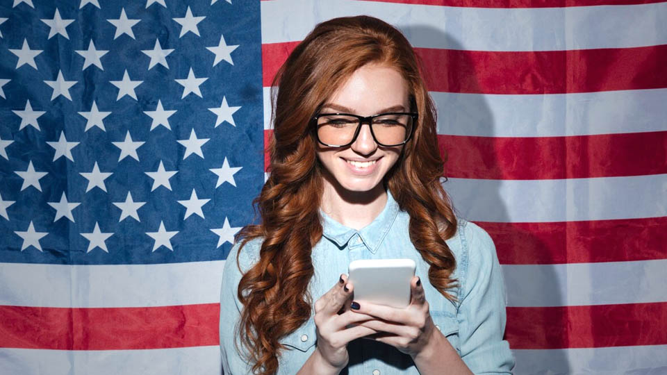 a cheerful woman looking at her phone with the usa flag in the background