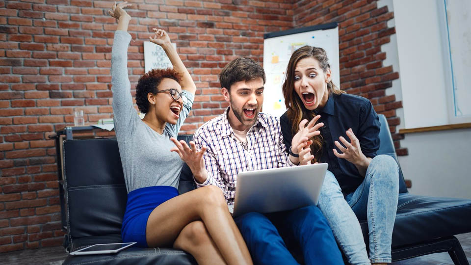 three excited people sitting on the sofa looking at a website on the laptop