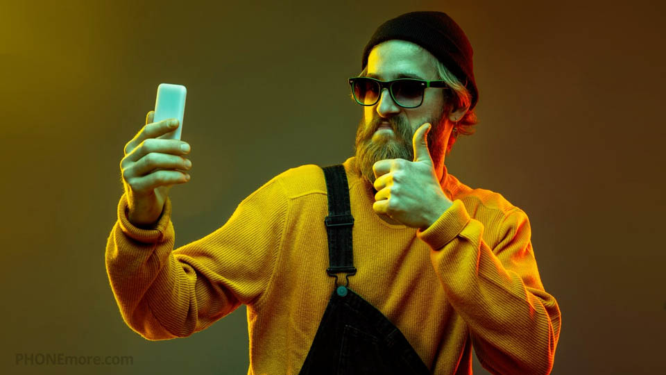 a man with a beard doing a thumbs-up with his hand and taking a selfie with his smartphone