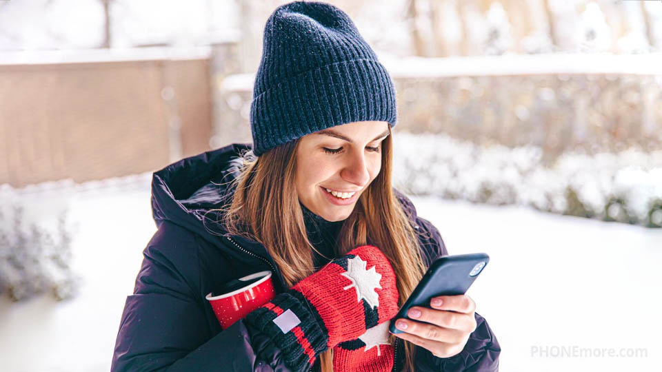 a happy woman in warm clothing and wearing a glove with the canadian flag looking at her smartphone