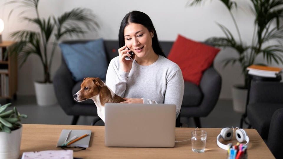 una mujer teletrabajando y hablando al móvil con su perro en casa en una mesa con ordenador portátil