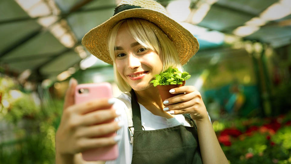 a woman holding a green plant and taking a selfie with her smartphone