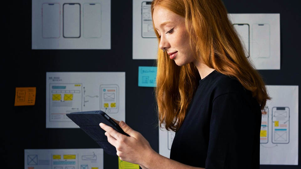 a red-haired woman looking at the tablet in an office with software development prototypes on the wall
