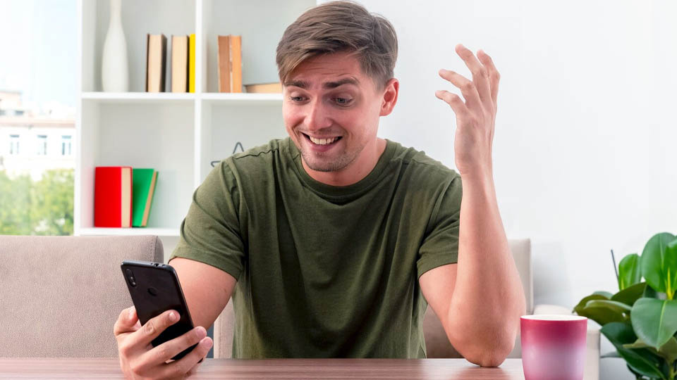 surprised young man sits at table holding and looking at phone inside living room