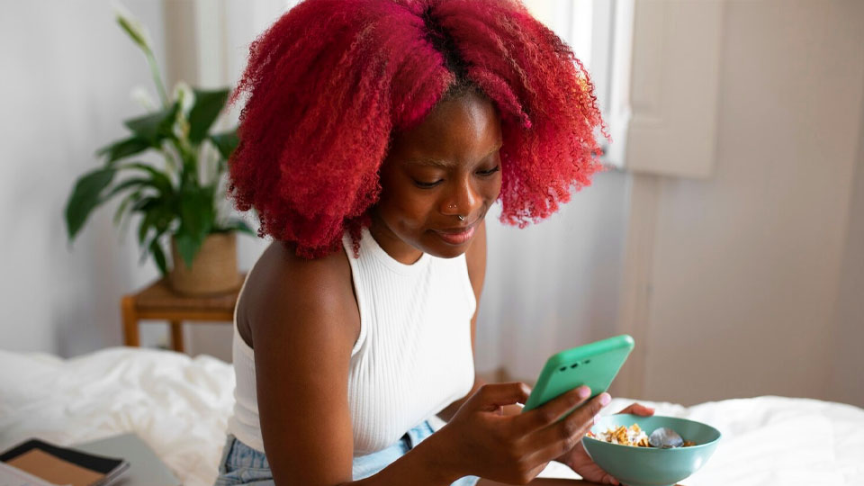 une femme assise sur le lit dans sa chambre sourit, son téléphone portable à la main