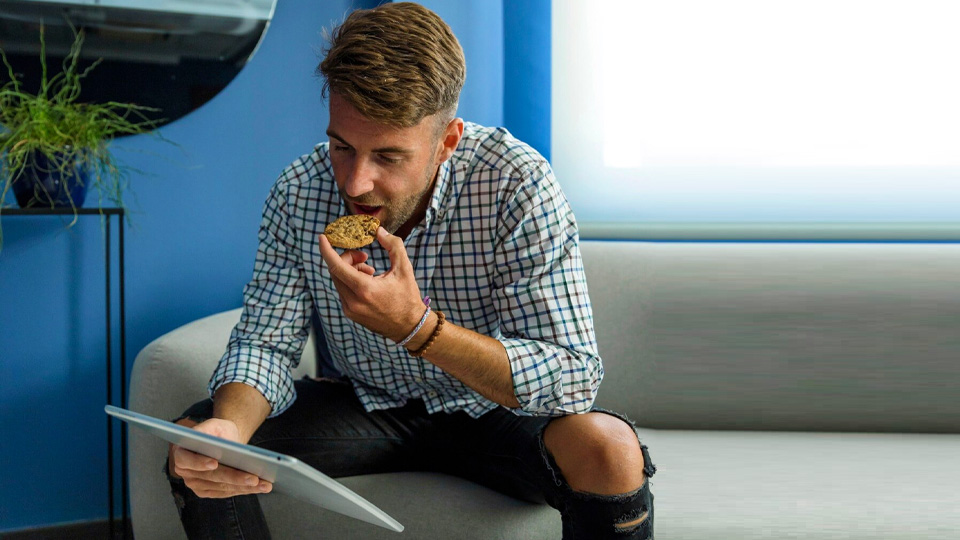 homme assis sur le canapé mange un biscuit en regardant sa tablette
